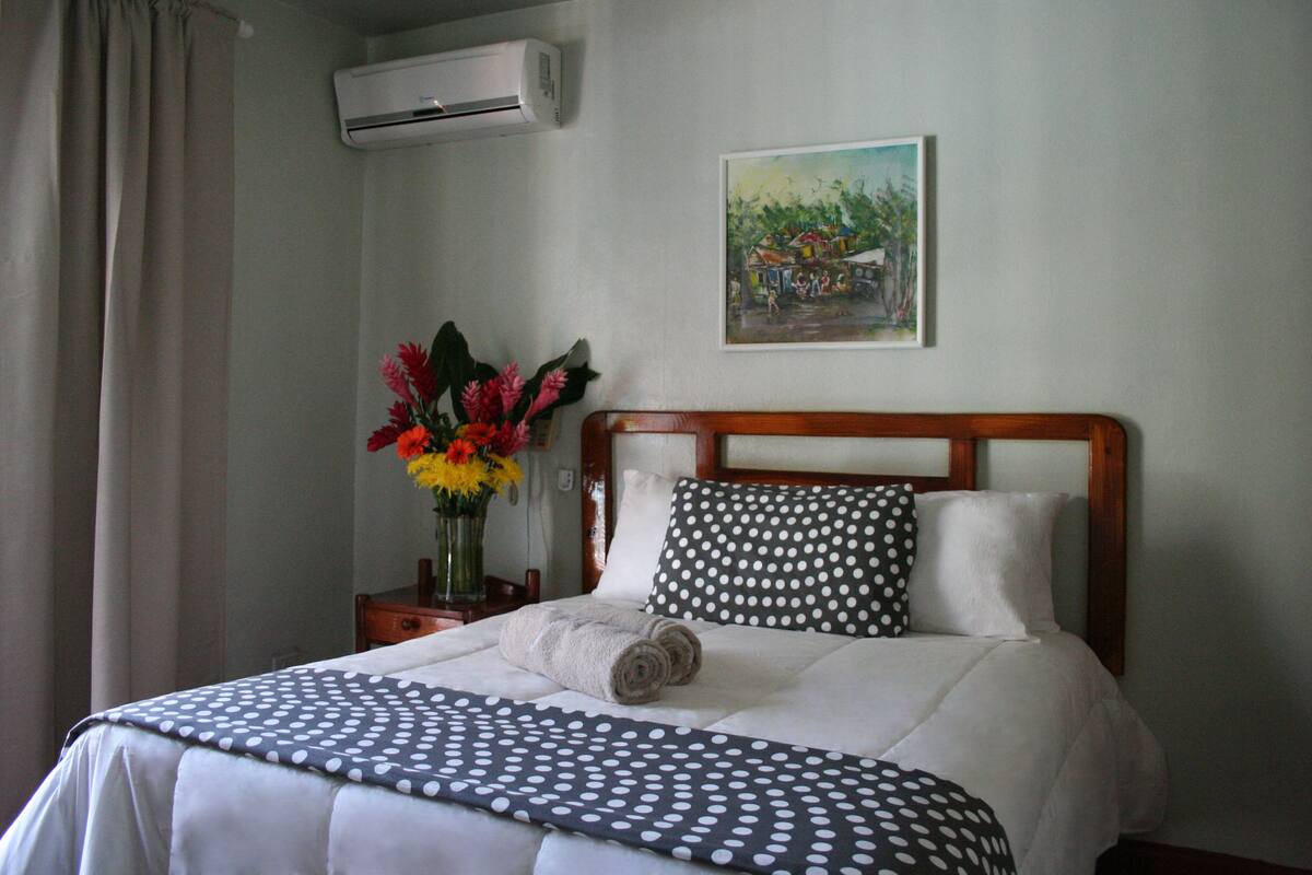 Une chambre d’hôtel avec un lit, une table de chevet ornée d’un vase de fleurs, un tableau au mur et un appareil de climatisation.
