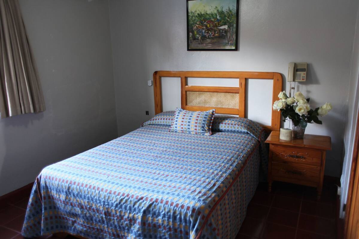 Une chambre à coucher avec un lit, une table de chevet ornée d’un vase de fleurs, un tableau au mur et un téléphone.