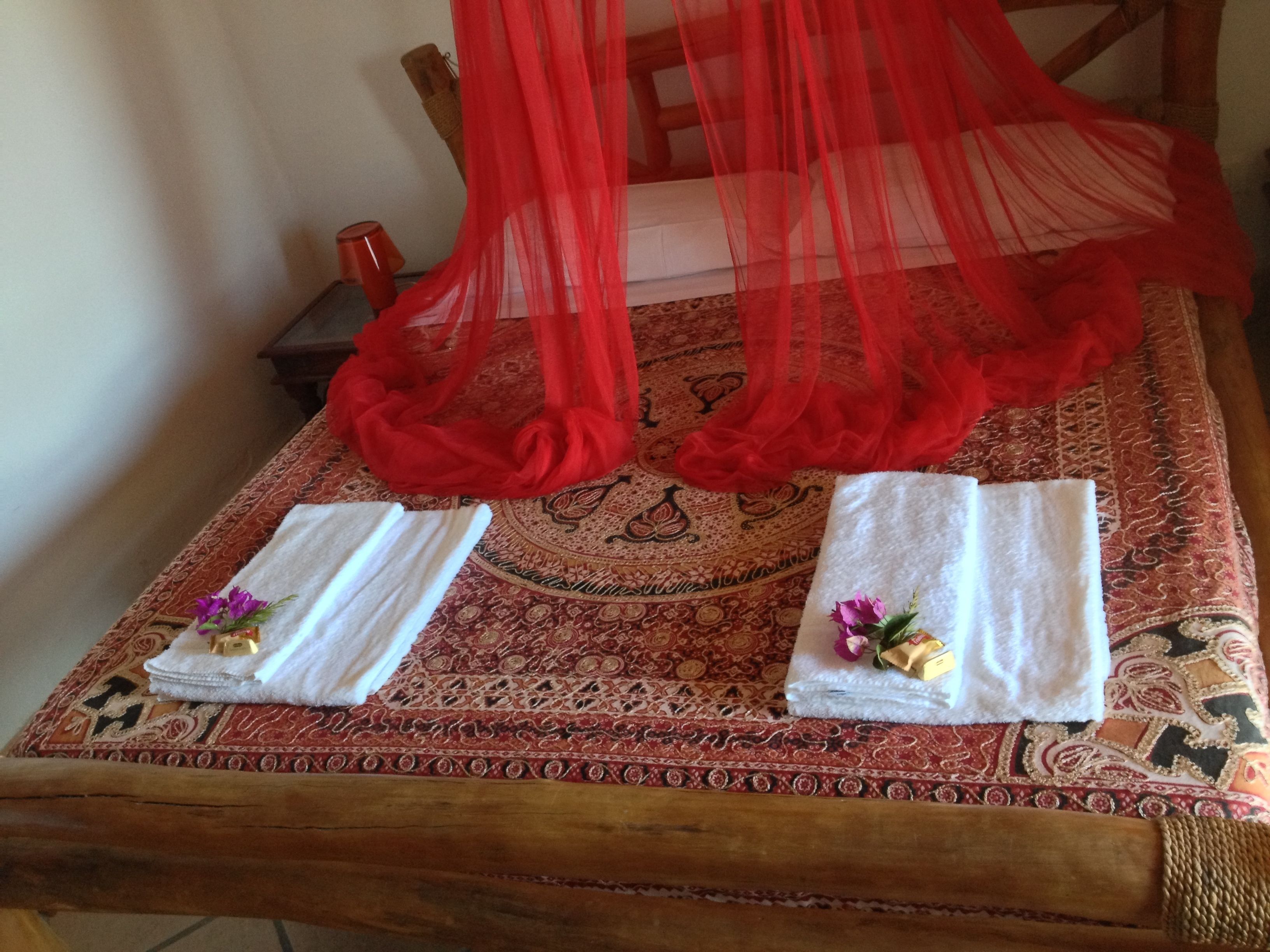 A bed with a red canopy, two towels with flowers, and a patterned rug.