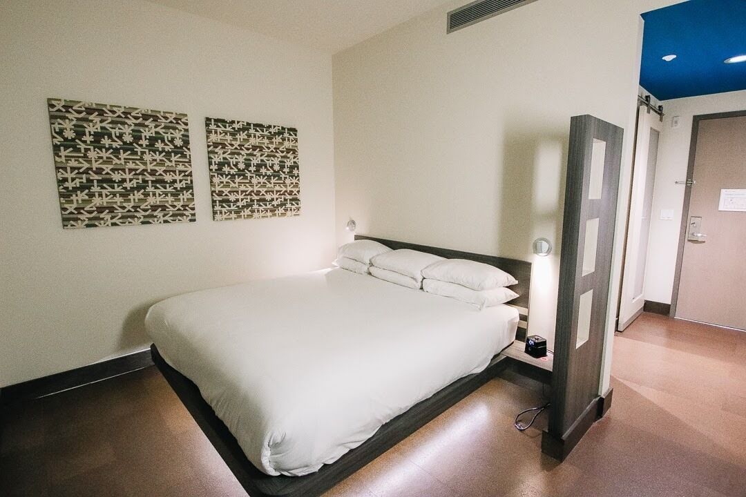 Camera d'albergo con un letto, due opere d'arte appese al muro, una porta e un interruttore della luce.