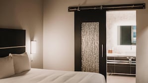 Ein Schlafzimmer mit Bett, Nachttisch, Wandlampe, einer Schiebetür zu einem Badezimmer und einem Fenster mit Ausblick.