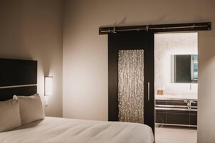 A bedroom with a bed, a nightstand, a wall-mounted light, a sliding door leading to a bathroom, and a window with a view.