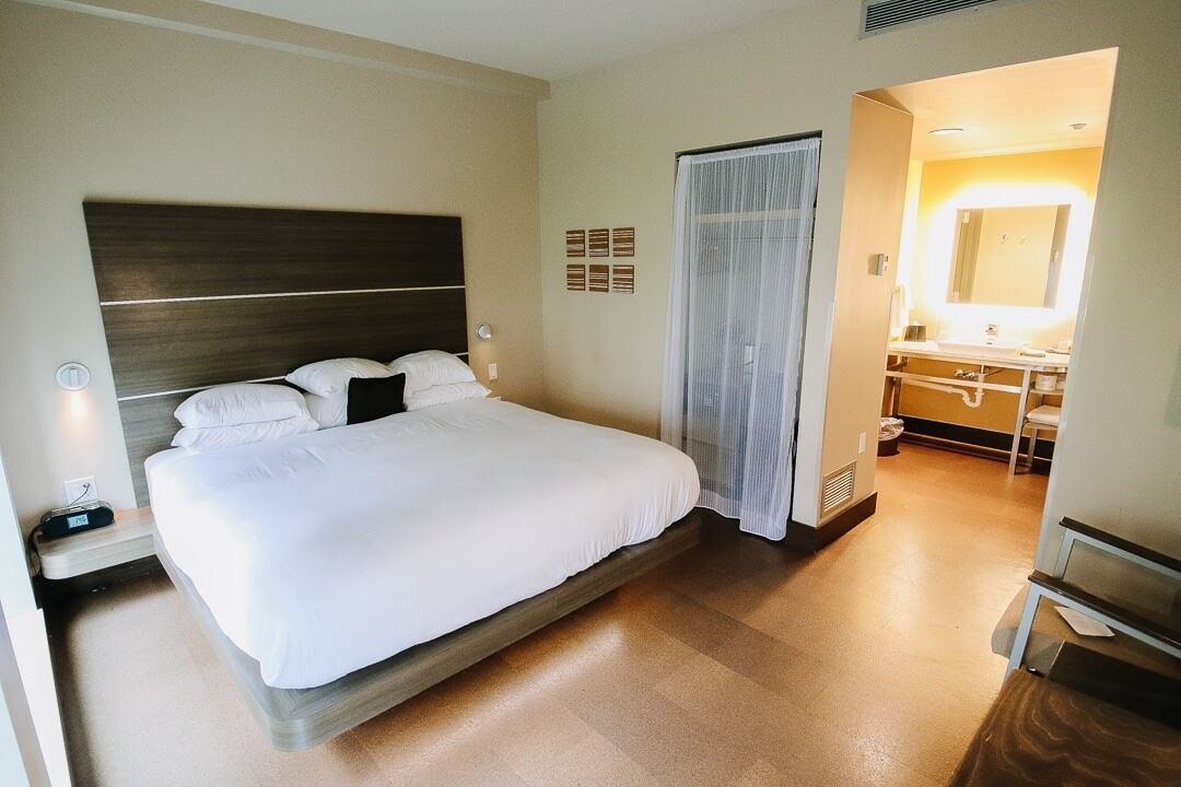 A hotel room with a large bed, bedside table, and a bathroom with a mirror and sink.
