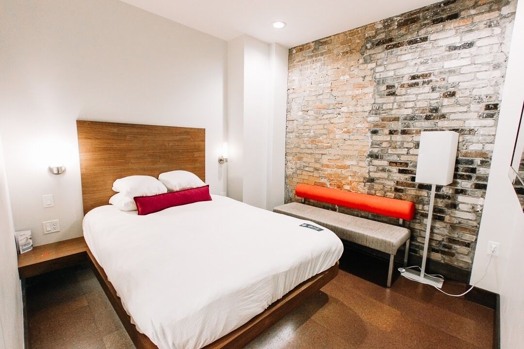 Et moderne hotelværelse med seng med trægavle, en væg med murstensdetaljer, en bænk og et natbord med lampe.
