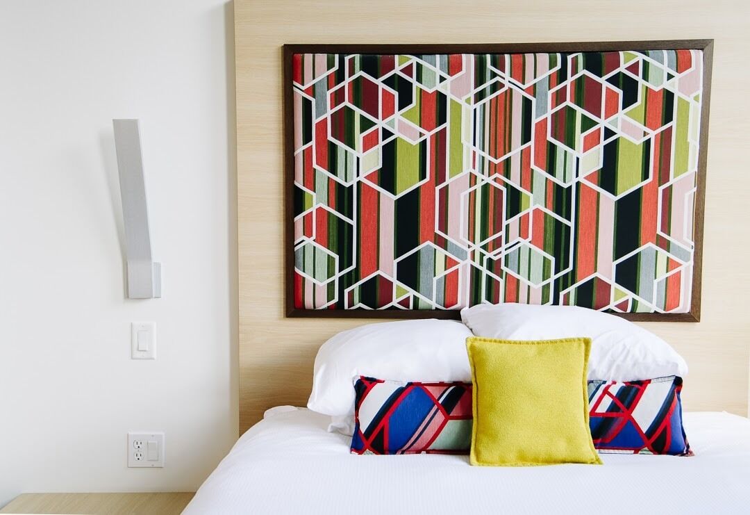 Een hotelkamer met een bed, wit beddengoed, kleurrijke geometrische wandkunst en een geel kussen.