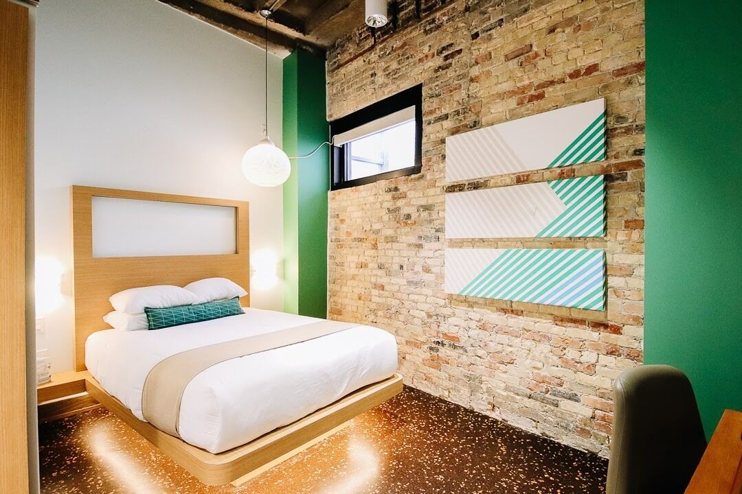 Une chambre d’hôtel moderne avec un mur en briques, un lit doté d’une tête de lit en bois, un mur d’accent vert et une fenêtre.