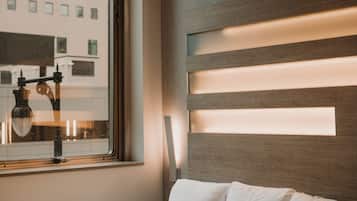 A hotel room with a large bed, a window with a view of buildings, and a wall with horizontal light panels.