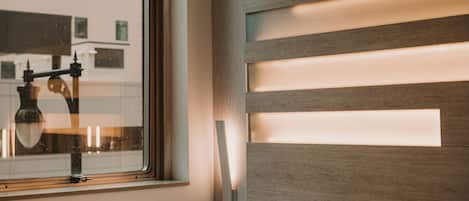 A hotel room with a large bed, a window with a view of buildings, and a wall with horizontal light panels.