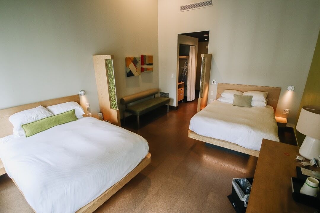 Habitación de hotel con dos camas, un banco, una mesita y un cubo de basura.