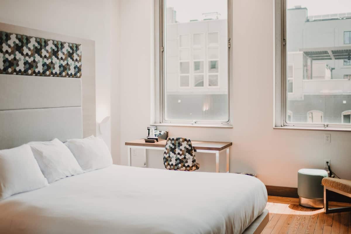 Ein Hotelzimmer mit Bett, Schreibtisch, Stuhl, Hocker und zwei Fenstern mit Blick auf Gebäude.