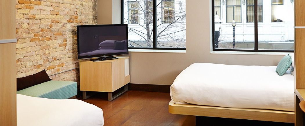 A hotel room with a brick wall, a bed, a TV on a wooden stand, and a window with a view of the outside.