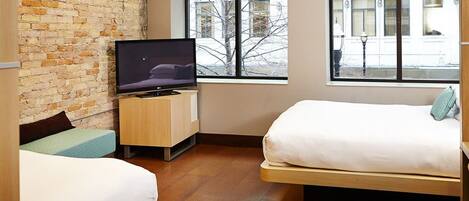A hotel room with a brick wall, a bed, a TV on a wooden stand, and a window with a view of the outside.