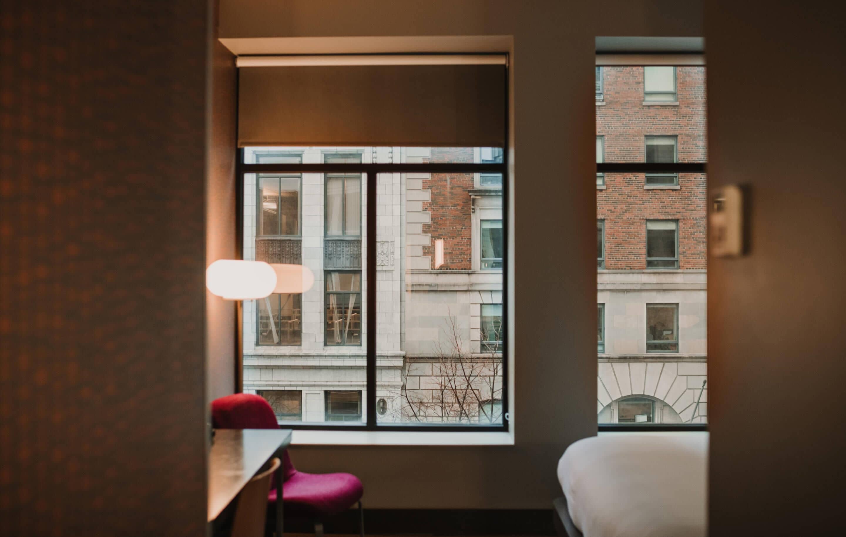 A hotel room with a large window, a desk, a chair, and a bed.