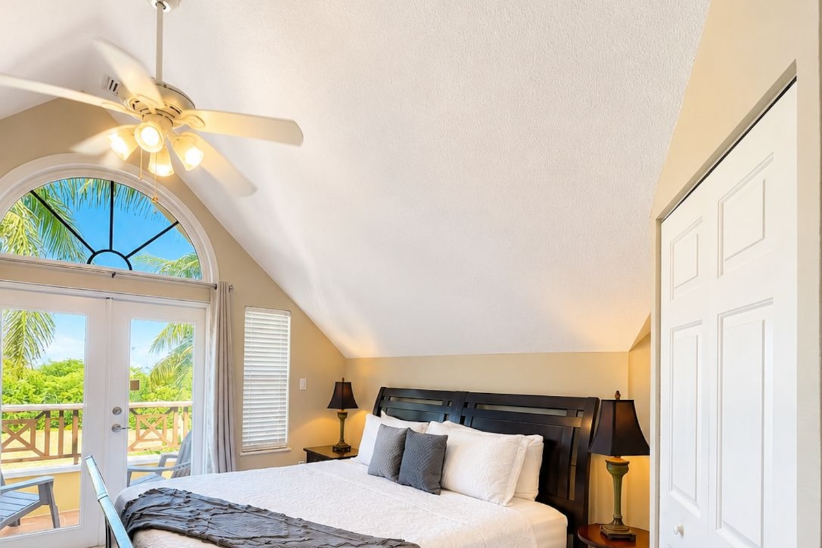 Ein Schlafzimmer mit einem Bett, einem Deckenventilator, einem Nachttisch, einer Lampe und Blick ins Freie.