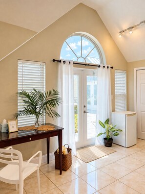 A room with a dining table, chairs, a potted plant, a small refrigerator, and a door with a view to the outside.