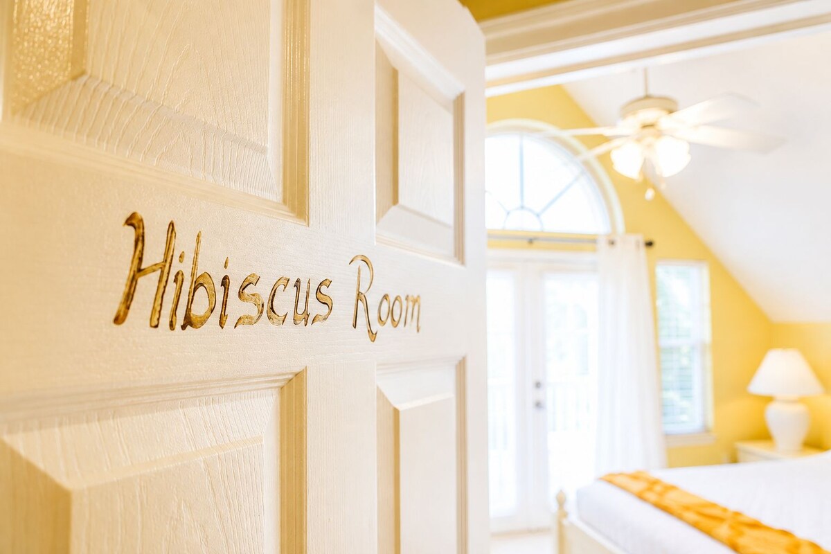 Ein Hotelzimmer mit einer T ür, auf der 'Hibiscus Room' steht, einem Bett, einem Deckenventilator und einem Fenster.