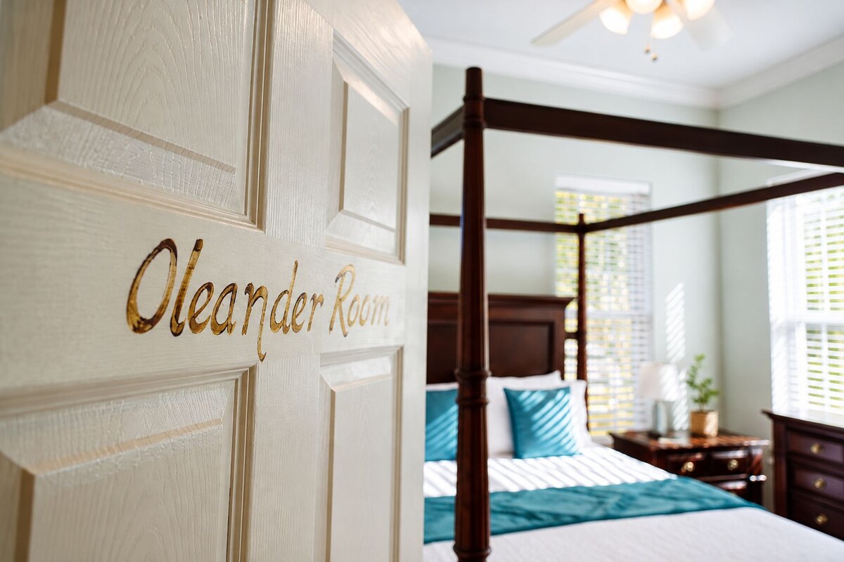Ein Schlafzimmer mit Himmelbett, einer Holztür mit der Aufschrift 'Oleander Room' und einem Fenster mit Jalousien.