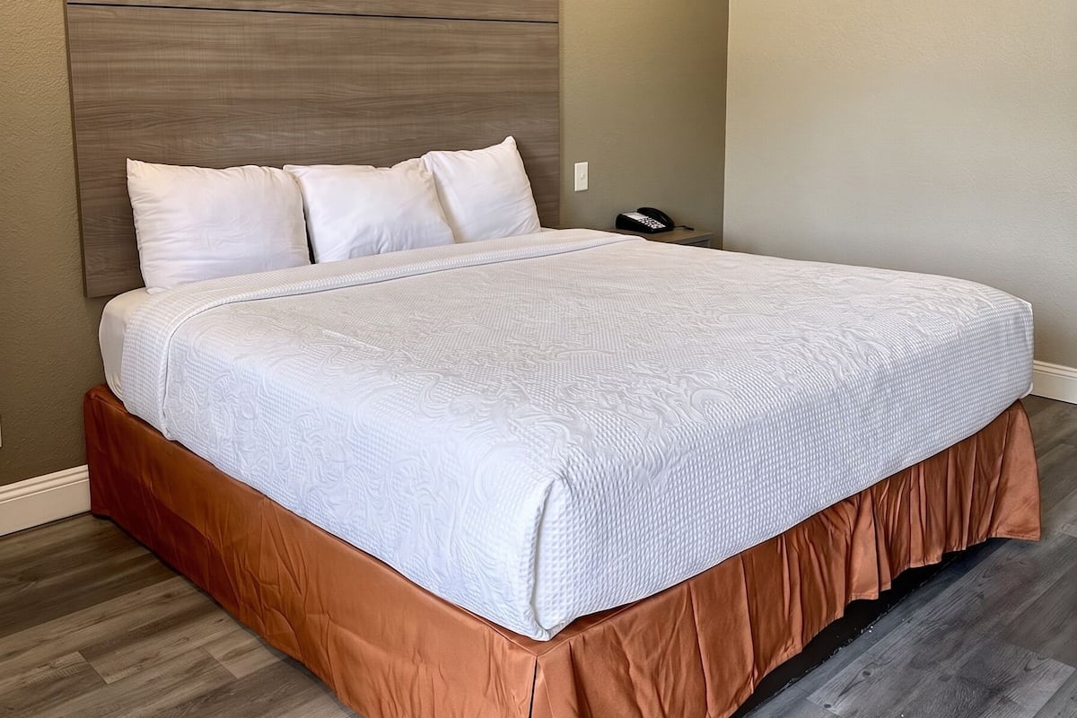 Uma cama bem arrumada com cabeceira de madeira, saia marrom e colcha branca em um quarto com piso de madeira e teto texturizado.