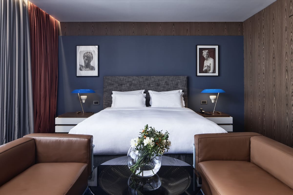 A modern hotel room with a large bed, two bedside tables with lamps, a small round table with a vase of flowers, and two brown leather chairs.