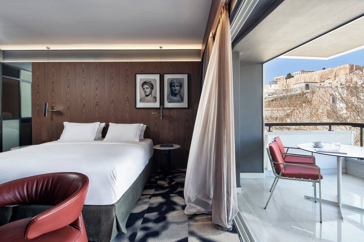 A modern hotel room with a large bed, a red chair, a small round table, and a balcony with a view of the city.
