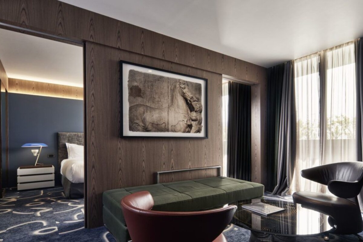 Ett modernt hotellrum med en grön soffa, ett inramat konstverk, en säng, en stol och utsikt över omgivningarna.