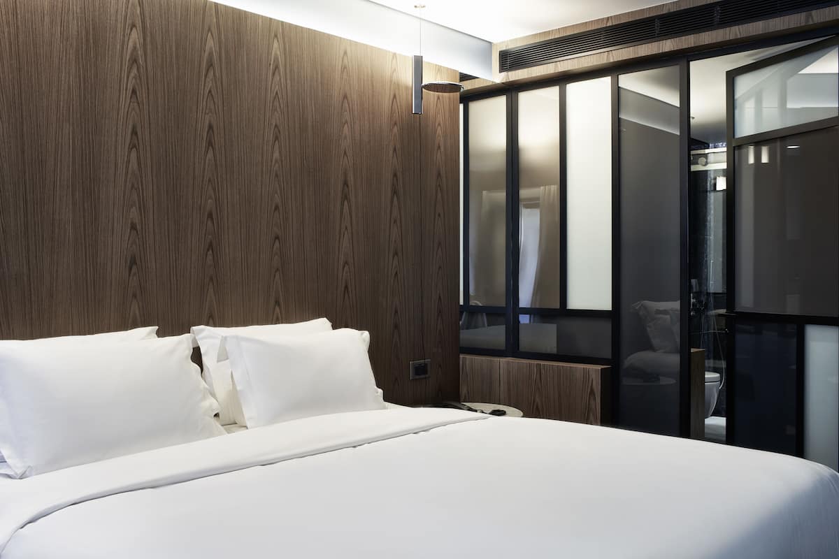 A modern hotel room with a large bed, wooden paneling, and a sliding glass door leading to a bathroom.