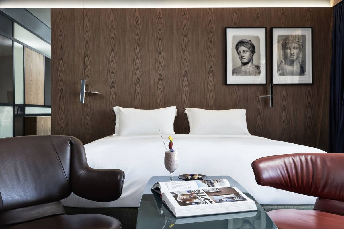 A modern hotel room with a large bed, two framed pictures on the wall, a glass coffee table with a magazine, and leather chairs.