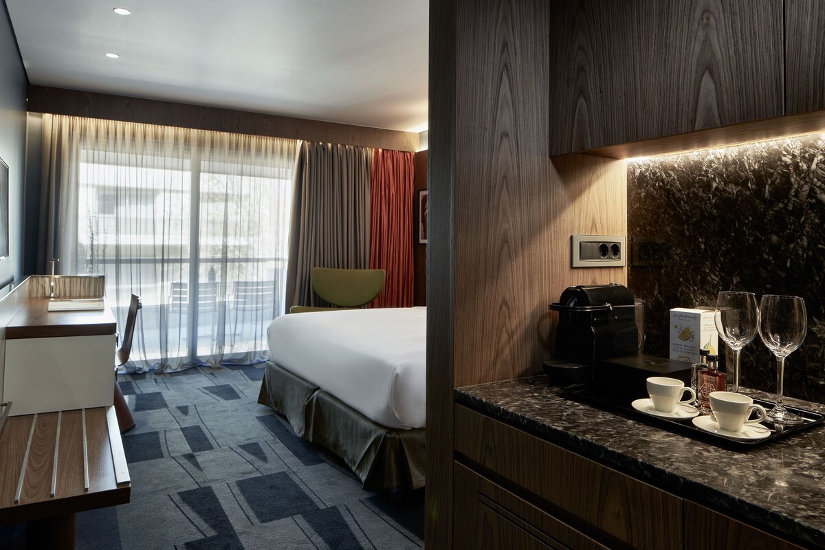 Ett hotellrum med en säng, ett skrivbord, en stol, en kaffemaskin och ett bricka med koppar och glas.