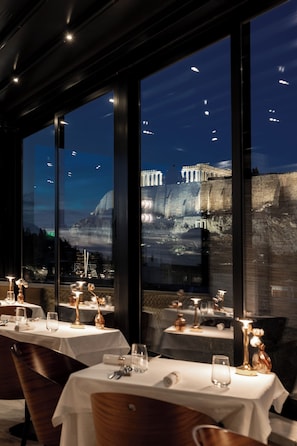 A restaurant with tables set for dining, large windows offering a view of a historic building at night. - AthensWas Design Hotel (Athens)