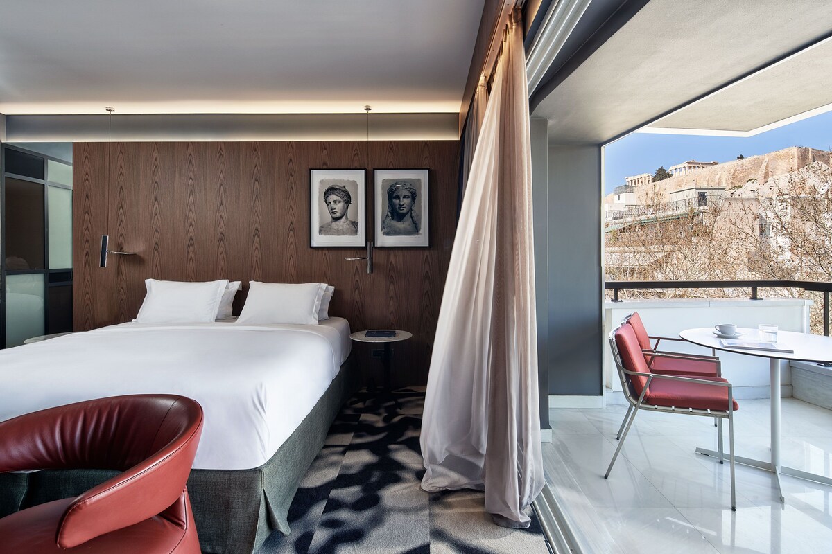 Ett modernt hotellrum med en stor säng, en röd stol, ett litet runt bord och en balkong med utsikt över en historisk byggnad.