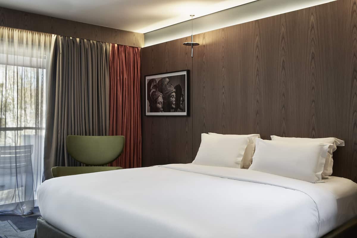 A hotel room with a large bed, a green chair, a framed picture on the wall, and a window with curtains.