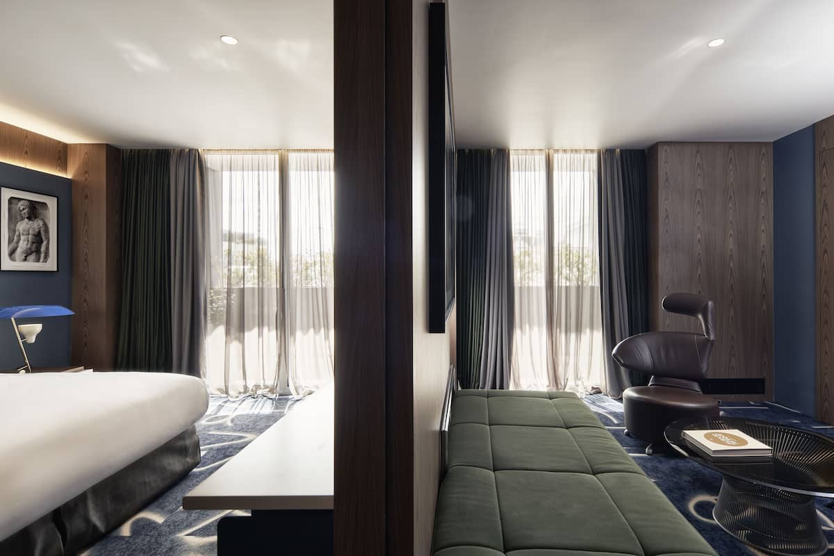 A modern hotel room with a bed, a sofa, a chair, a table, and a large window with curtains.