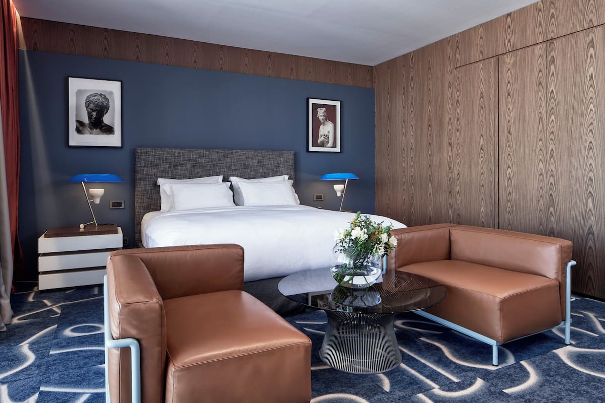 A hotel room with a large bed, two side tables with lamps, a brown leather sofa, a glass coffee table, and a blue patterned carpet.