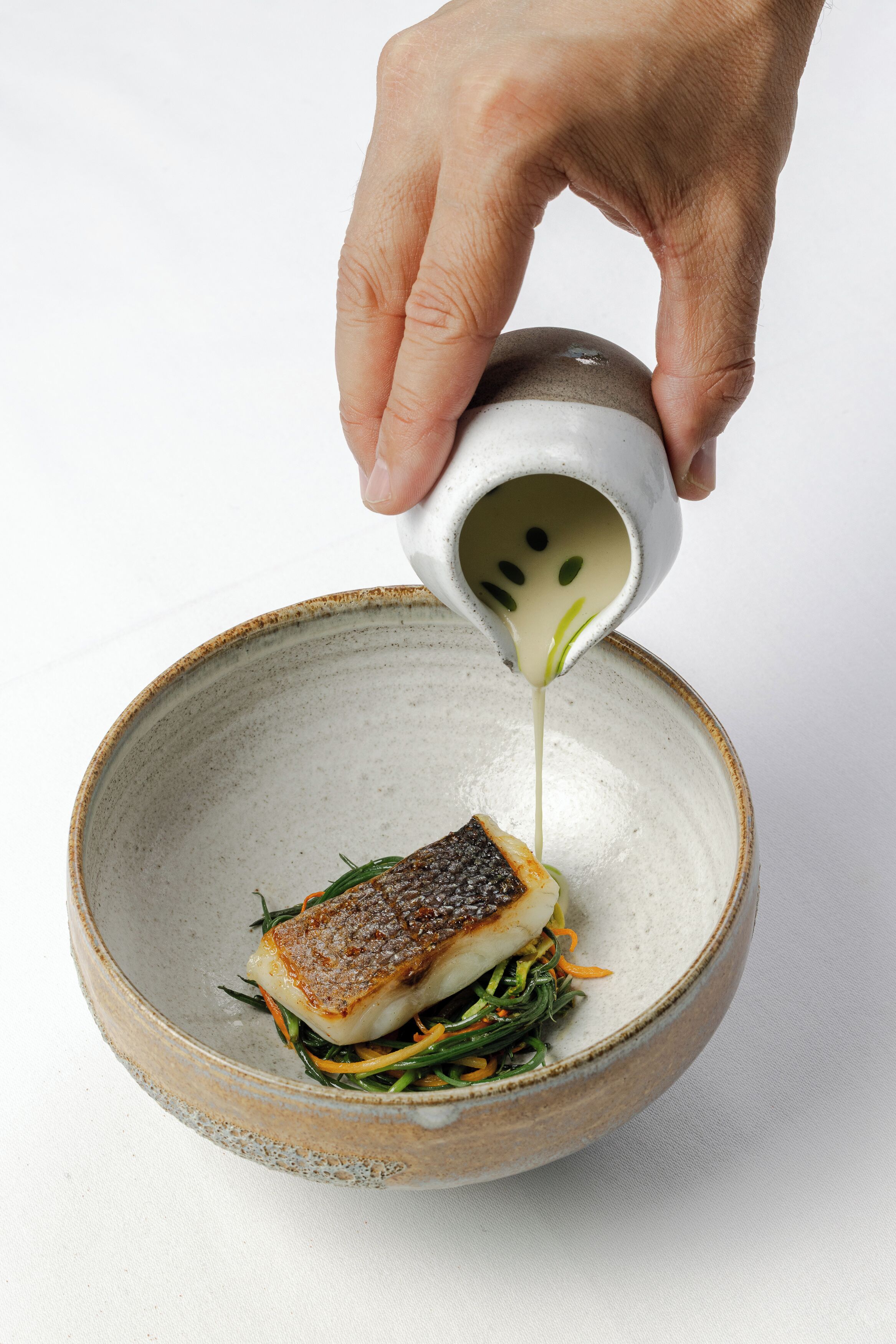 Une main verse une sauce sur un plat de poisson dans un bol en céramique.