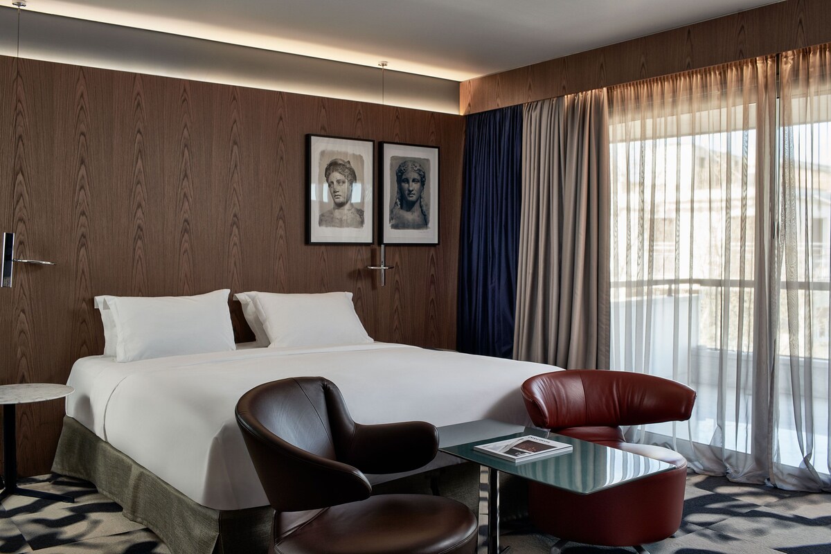 Ett hotellrum med en säng, två stolar, ett litet bord och ett fönster med gardiner.