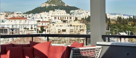 A balcony with a red sofa, a wicker chair, and a small table with a plant. A view of a cityscape with buildings and a hill is visible in the background.