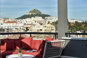 A balcony with a red sofa, a wicker chair, and a small table with a plant. A view of a cityscape with buildings and a hill is visible in the background.