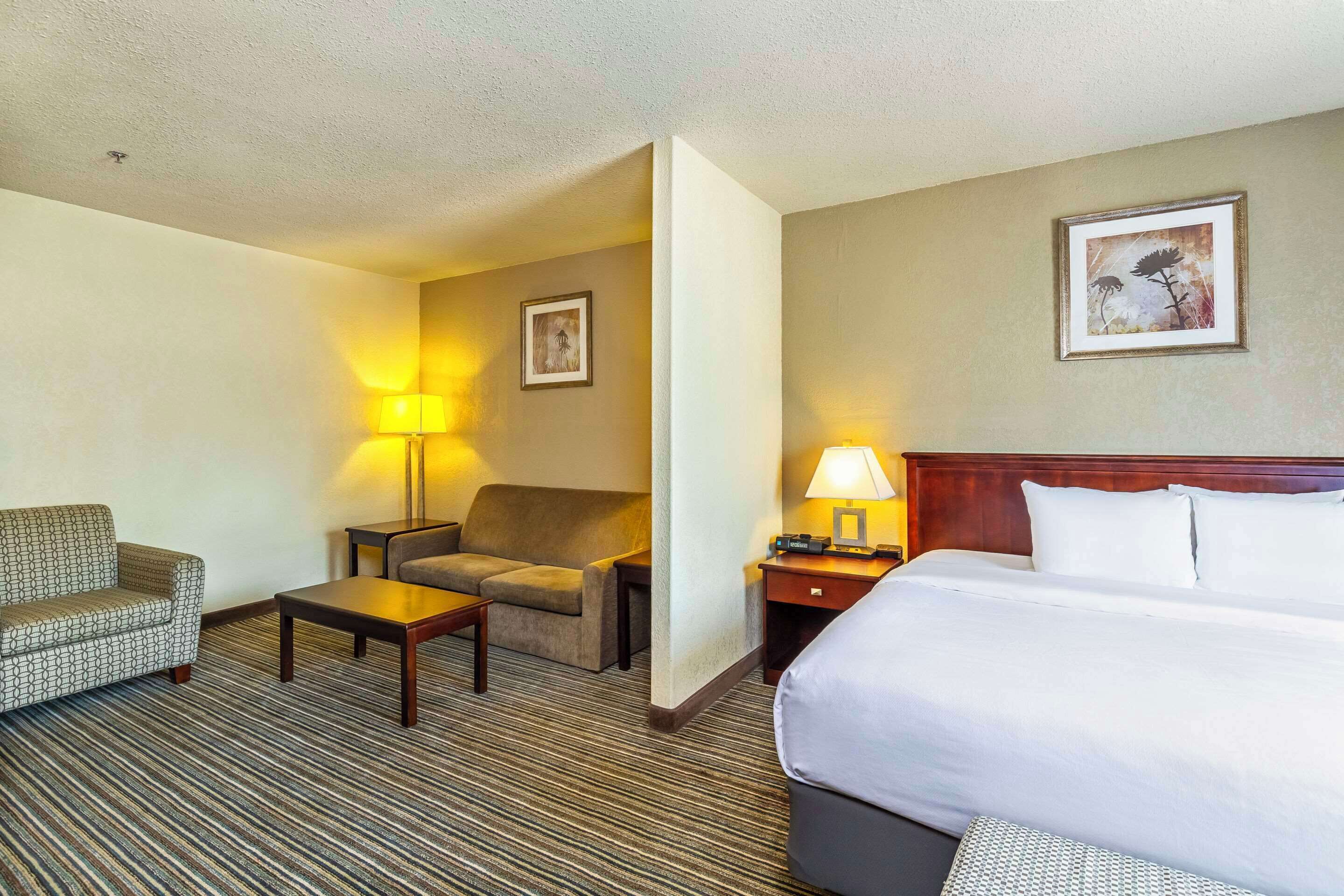 Habitación de hotel con una cama grande, dos sillones, una mesita y una mesita de noche con lámpara.