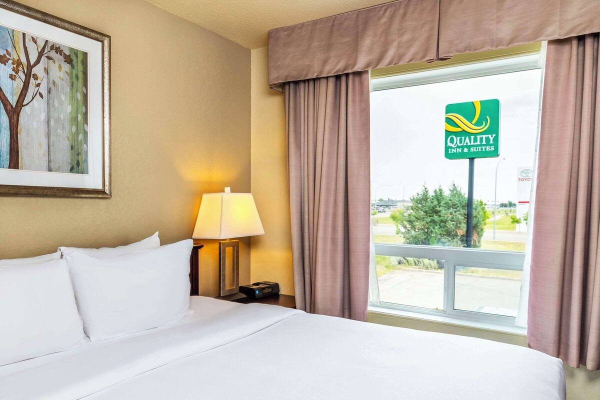 A hotel room with a bed, a lamp, a framed picture, and a window with a view of the outside.