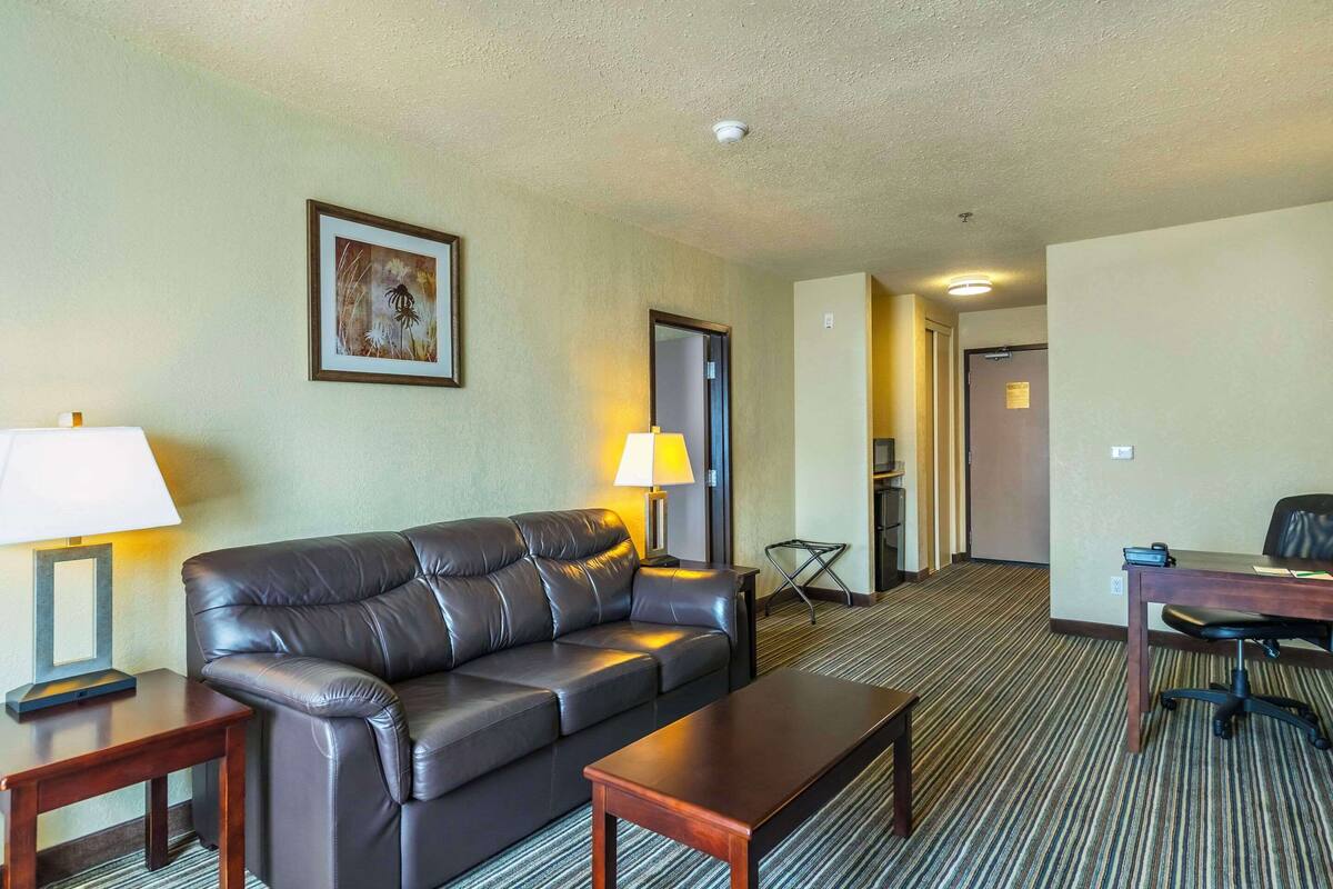A hotel room with a leather sofa, a wooden coffee table, a desk with a chair, a lamp, and a framed picture on the wall.
