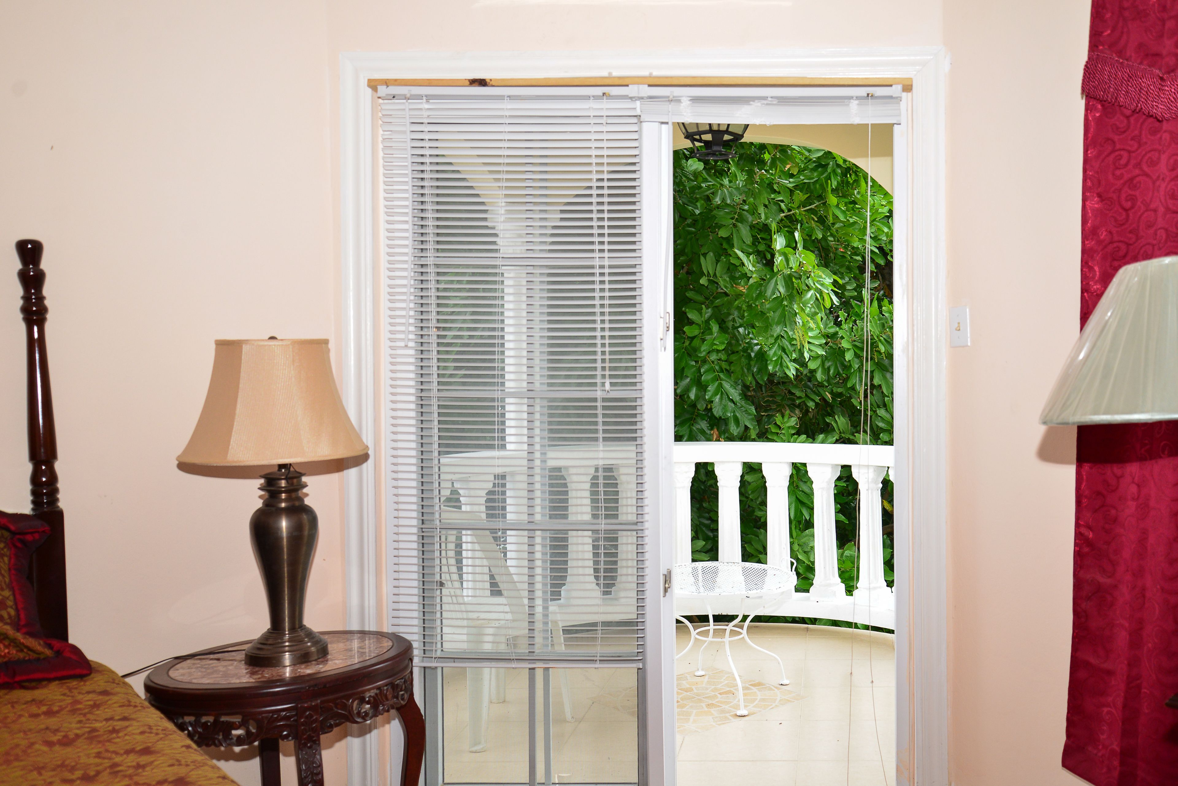 A room with a wooden table and a lamp, a white railing, and a view of greenery outside.