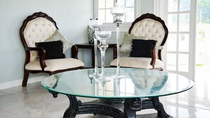 A round glass table with three wine glasses, two upholstered chairs with dark wood frames and white cushions, and a window with white panes in the background. - Khastle Khodero (Montego Bay)