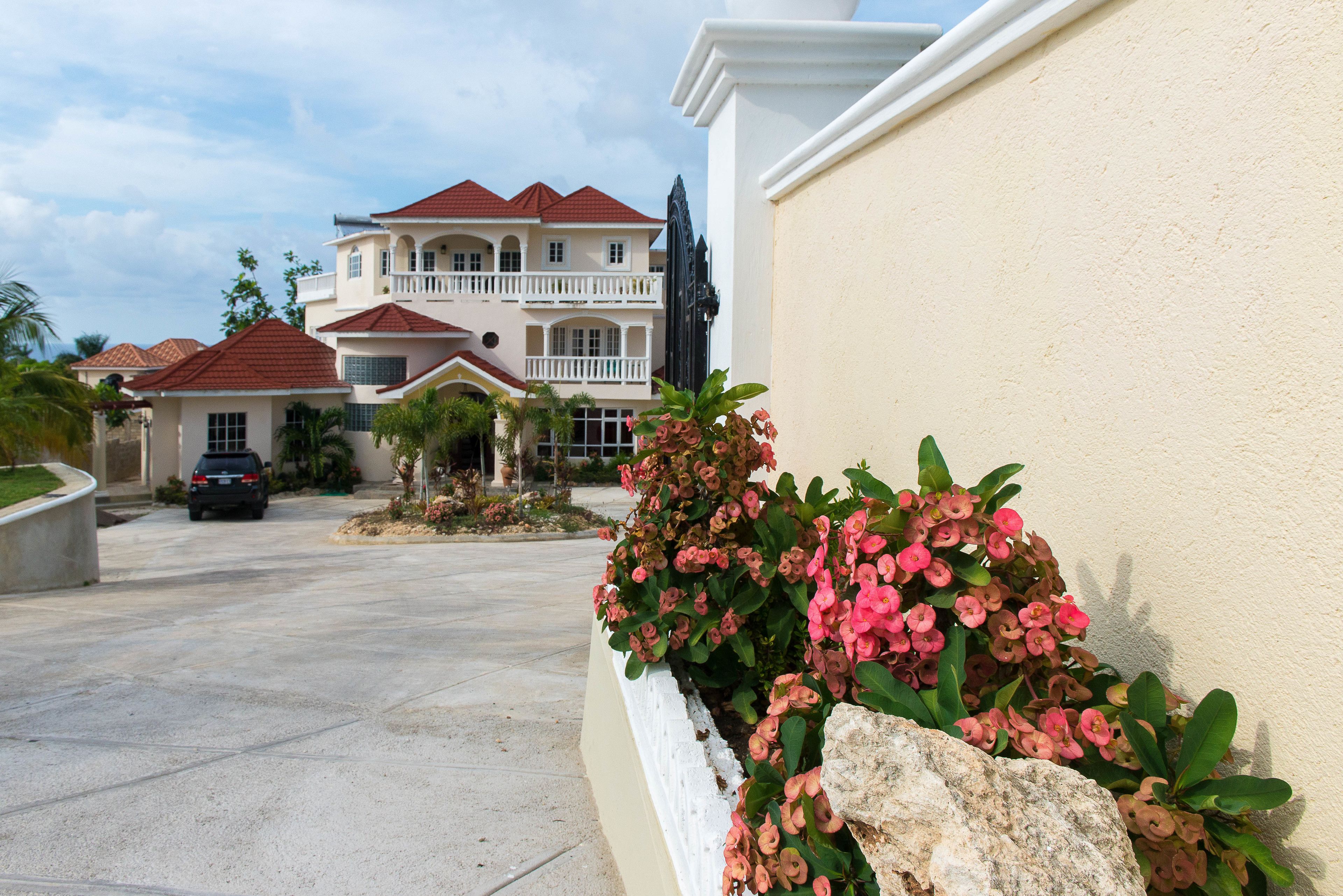 A residential street with a large house featuring a balcony, a driveway, and a flower bed with pink flowers.