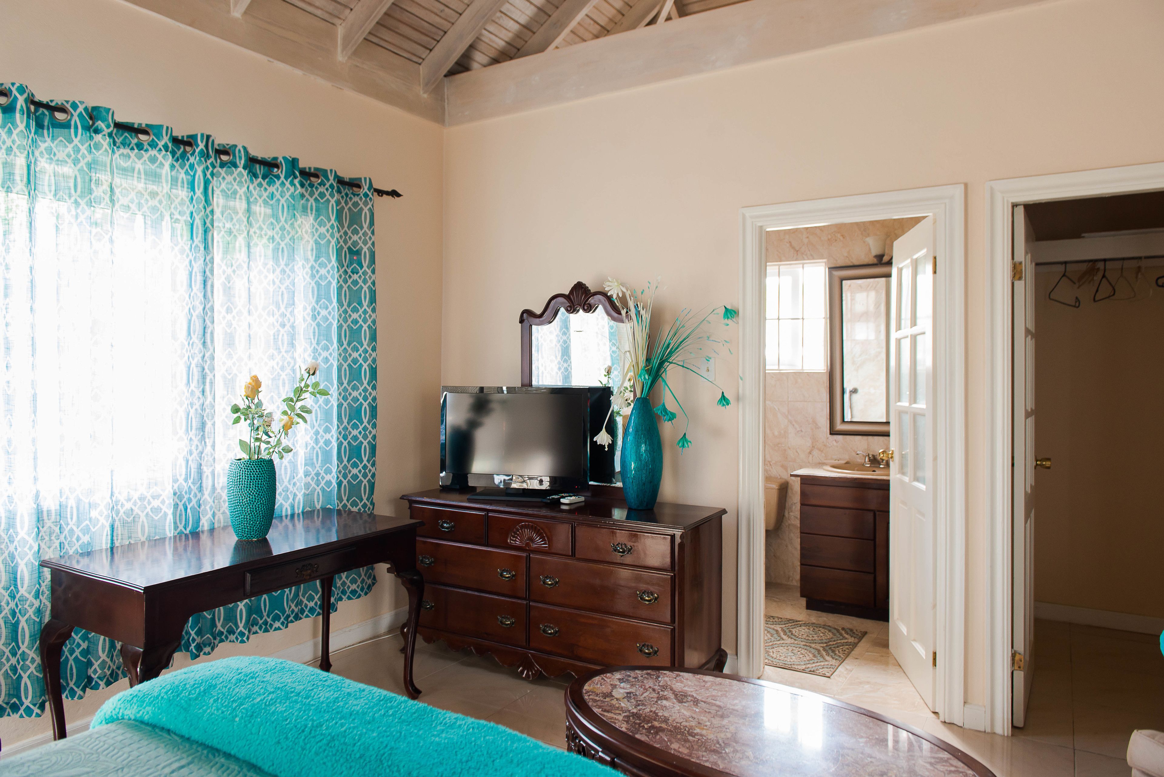 A room with a wooden dresser, a television, a vase with flowers, a bed, and a bathroom visible through an open door.