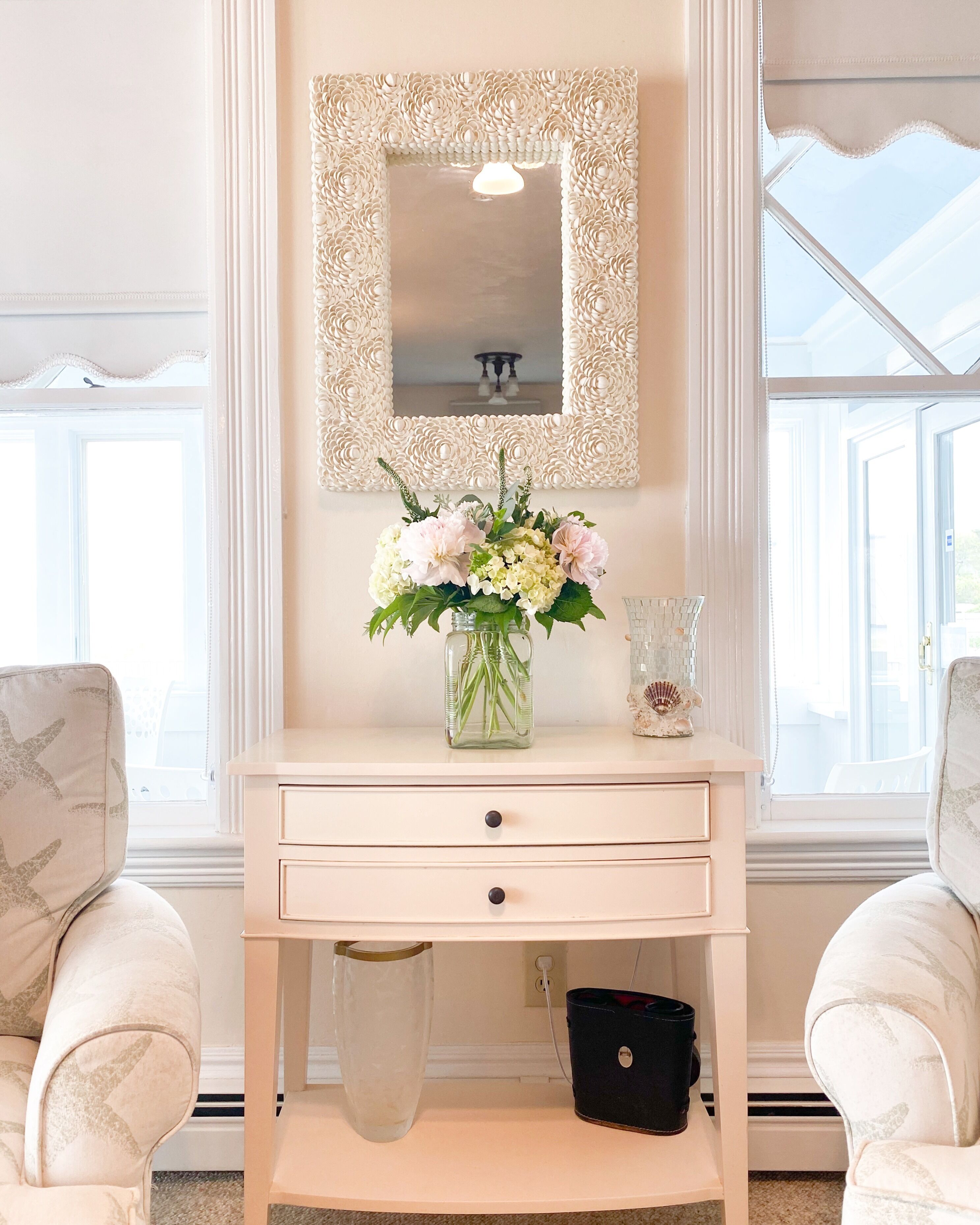 Ein Zimmer mit einem hellen Schminktisch, einem Spiegel mit floralem Rahmen, einer Vase mit Blumen und zwei Stühlen.