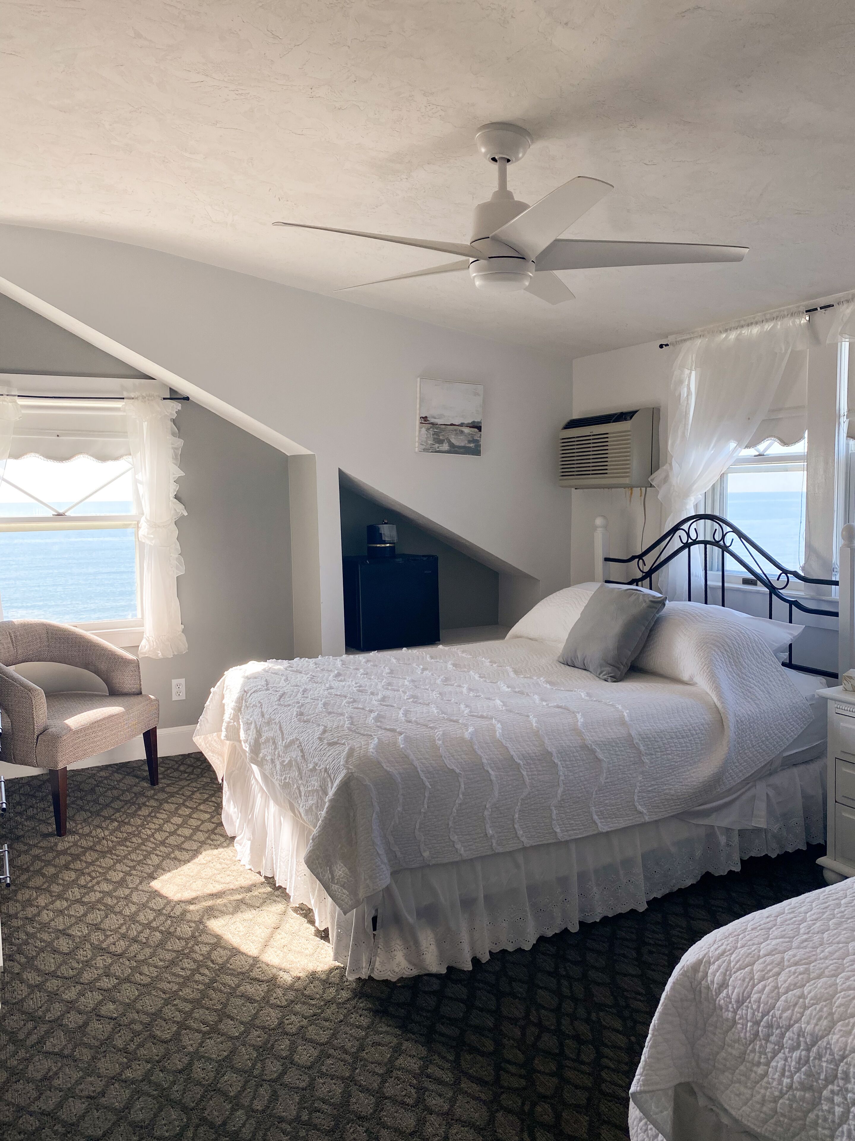 A bedroom with a bed, a chair, a ceiling fan, a window with curtains, and a view of the sea.