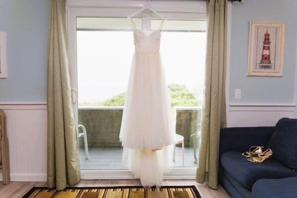 A room with a white dress hanging in front of a sliding glass door, a dark blue sofa, and a framed picture of a lighthouse on the wall. - By the Sea Guests Bed & Breakfast & Suites (Dennis Port)
