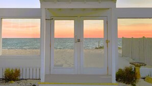 Een strandhuis met een witte gevel, twee grote ramen en uitzicht op de oceaan bij zonsondergang.