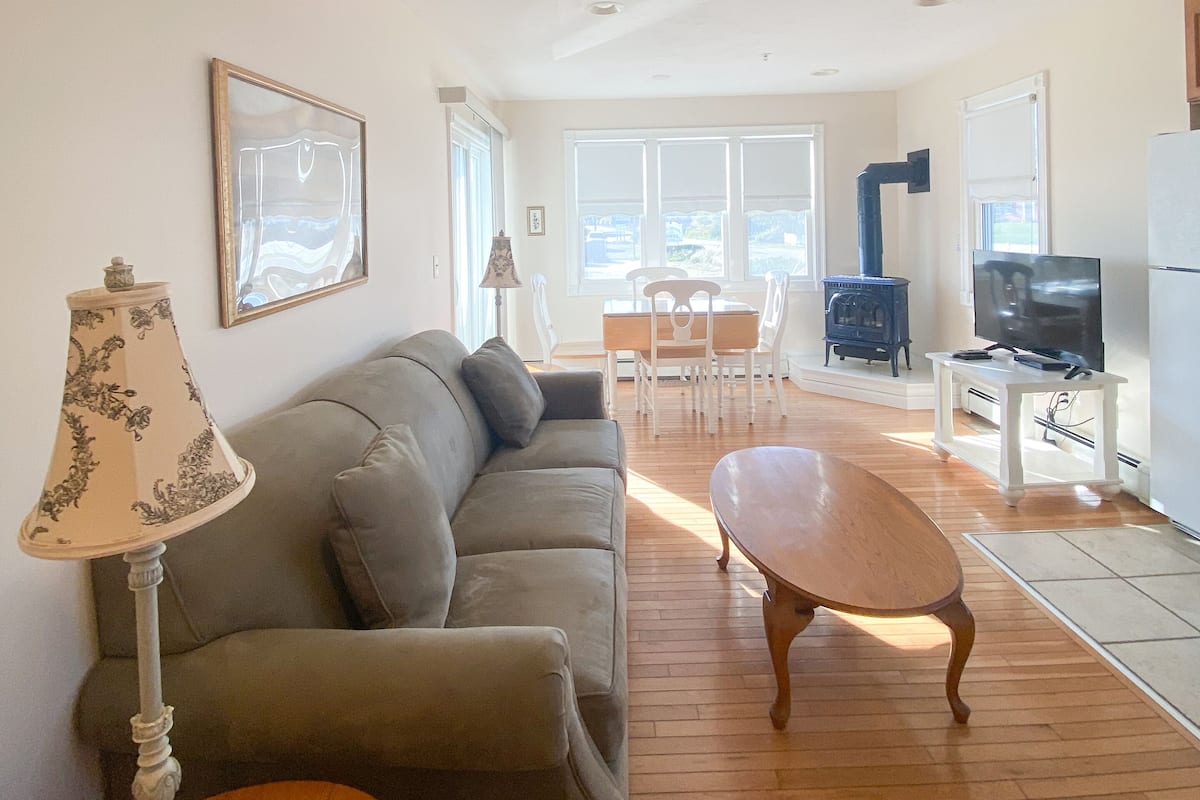 Een woonkamer met een bank, een houten salontafel, een lamp en een tv-meubel. Er is een eethoek met een tafel en stoelen, en een keuken met een fornuis en een koelkast.