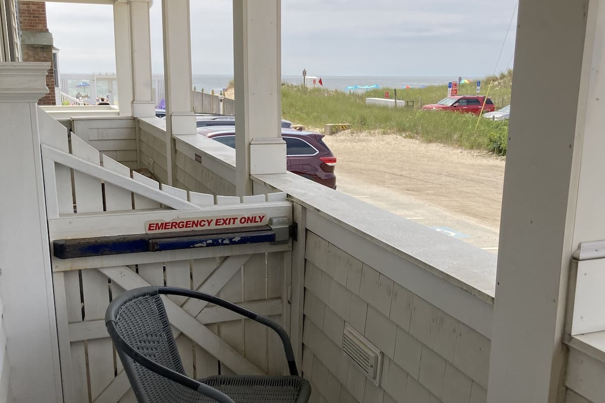 Een overdekt buitengebied met twee rieten stoelen, een witte leuning en uitzicht op het strand en geparkeerde auto's.